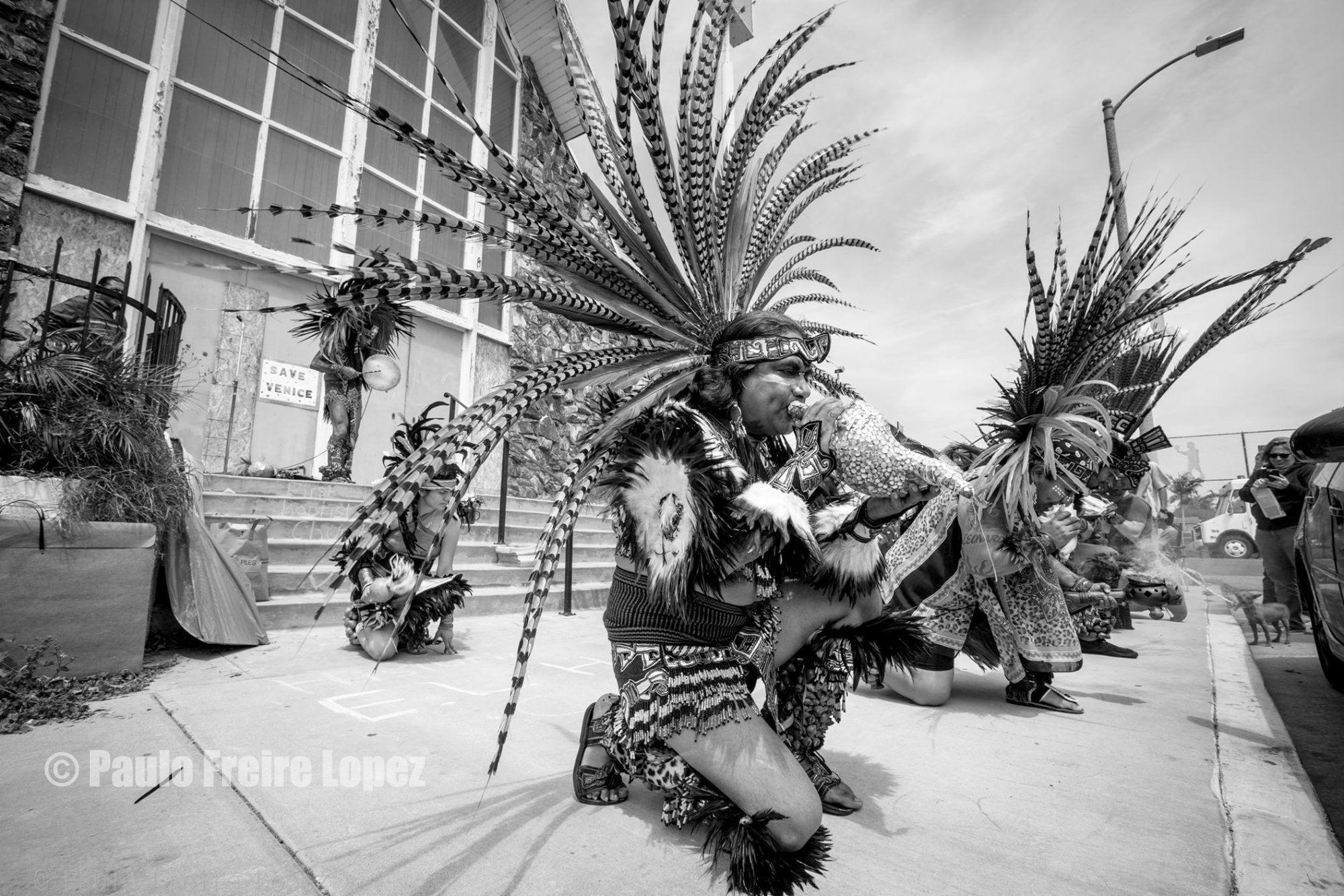 Danza Azteca dancers in feathered regalia kneel outside a church in Oakwood, with a hand-painted Save Venice sign by the door