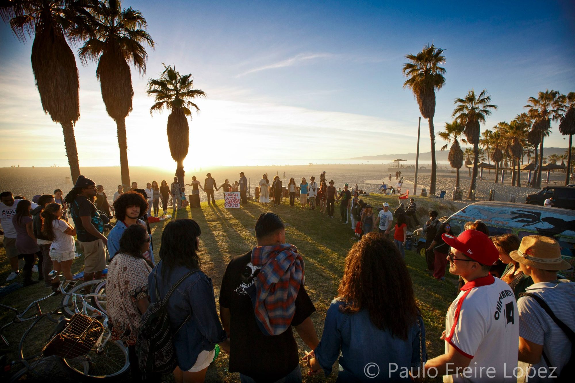 A community circle holding hands at sunset on the grass above Venice Beach