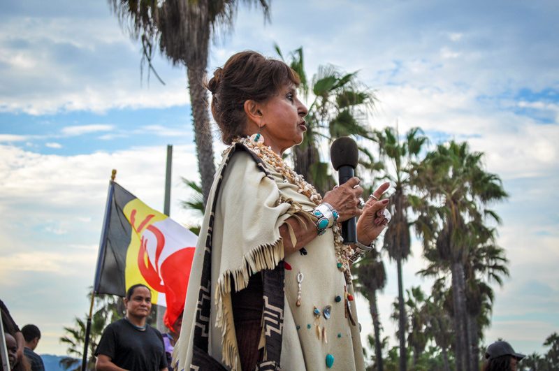 A woman in a fringed shawl speaks into a microphone at a gathering under the palm trees near the beach
